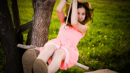A teenage girl in a summer hat swings on a swing in the forest. Walk on a sunny summer dayの写真素材