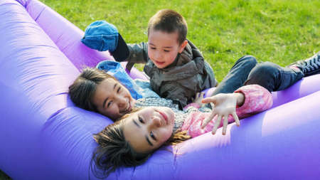 Happy little brother and two sisters lie on an inflatable chair against the background of green grassの写真素材