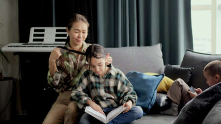 Mom braids her daughters hair in the morning, sitting on the sofa in the living room. little daughter reading a bookの写真素材