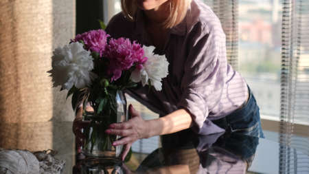 A young woman standing in a modern cozy living room puts a vase with a bouquet of beautiful fresh...