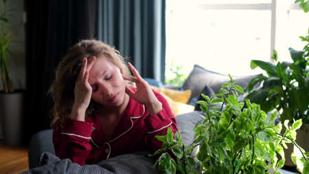 A sad young woman is suffering from a severe headache. Curly woman sits on the couch and grimaces...
