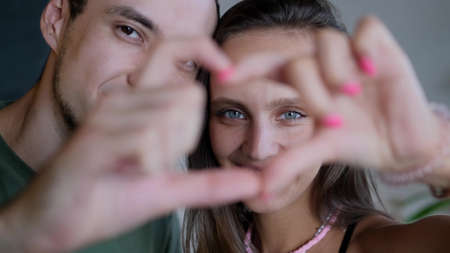 Close up portrait happy couple faces looking through joined fingers making heart shape. St Valentines Day celebration, romantic relationships, just married spouses, sincere feelings and love conceptの写真素材