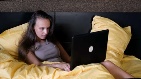 Beautiful woman is using her laptop while lying on the bed. She is typing a message on the laptop keyboard.の写真素材