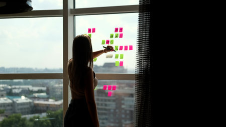 Concentrated caucasian woman writes notes on colorful paper stickers placed on a large window....