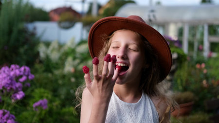 Cute little girl in a hat funny eats red raspberries on her fingers in the summer in the gardenの写真素材