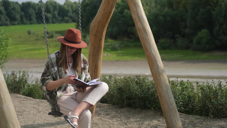 Satisfied smiling girl in a hat sits on a swing in the park and writes in a notebook. Outdoor portrait of a beautiful young womanの写真素材