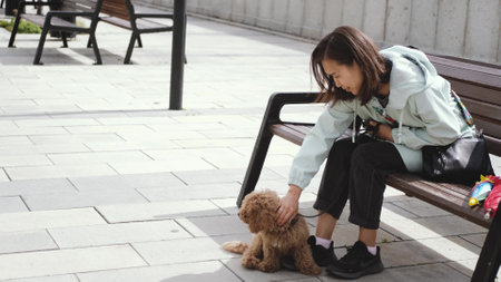 Pretty young Asian woman in a jacket is resting on a bench while walking her dog. Happy woman and her dog on a daytime walk.の写真素材