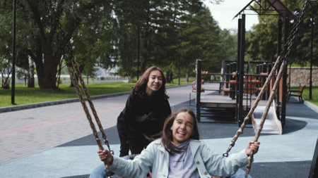 Two happy sisters are swinging. A cute little girl is pushing the swing, swinging it.