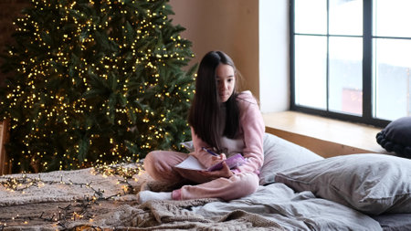 A cute little girl writes letters to Santa Claus, sitting on a cozy bed in front of a Christmas tree...