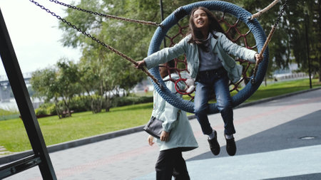 Happy mom and daughter are resting in the city park. Mom swings a laughing girl on a swing, pushes the swing.の写真素材