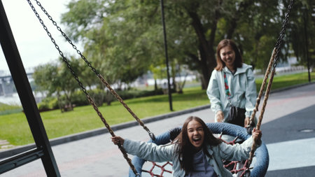 Happy mom and daughter are resting in the city park. Mom swings a laughing girl on a swing, pushes...