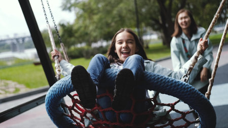 Happy mom and daughter are resting in the city park. Mom swings a laughing girl on a swing, pushes the swing.の写真素材