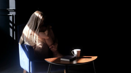 A depressed young beautiful woman sits alone in an armchair in the dark, hugs her knees and is sad. Loneliness sad woman thinks about grief, problems of life. Negative emotions andの写真素材