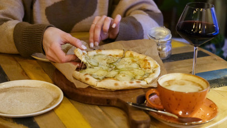 A womans hand takes a piece of hot pizza with pear and cheese, sitting in a cafe in the evening.