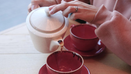 A woman in a pink sweatshirt pours hot tea into a ceramic cup in a cafe to keep warm in the cool...