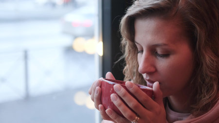 A woman in a pink hoodie holds a red ceramic cup with a hot drink and keeps warm in the cool evening...