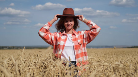 A young woman in a brown hat is enjoying a beautiful sunny warm day in a wheat field. View of a happy woman who smiles and poses for the camera against the background of golden wheat with ripe ears.の写真素材