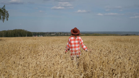 A young woman in a brown hat is enjoying a beautiful sunny warm day in a wheat field. View of a happy woman who runs, has fun and spins against the background of golden wheat with ripe ears.の写真素材