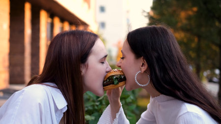 Two teenage friends biting the same hamburger at the same time, two friends eating junk food on the...
