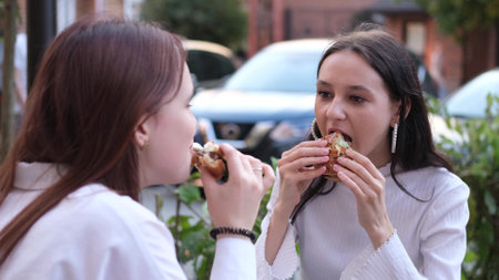 Two teenage girls are eating hamburgers and talking while sitting on a bench outside on a warm summer day.の写真素材