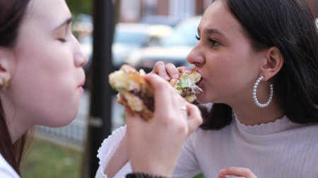 Two teenage friends biting the same hamburger at the same time, two friends eating junk food on the...
