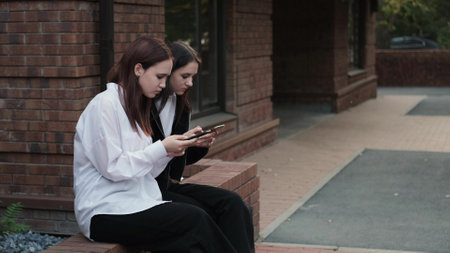 Cute teenage girls sitting on a street bench and looking at smartphones - friends hanging out outdoorsの写真素材