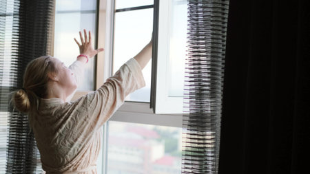 A young attractive woman in home clothes opens the window and enjoys the fresh air. The woman woke up and rejoices in a new day.の写真素材
