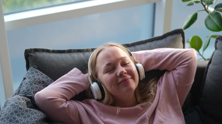 A serene young woman is relaxing on a comfortable sofa listening to music on headphones. Pretty woman likes to listen to relaxing music, sing without feeling stress at home.の写真素材