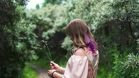 A young woman enjoys the beautiful weather, enjoys a wild flower outdoors with a smile. The woman is wearing a pink long boho dress.の写真素材