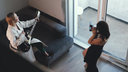 Two best friends have fun together. Young beautiful woman plays the guitar and sings a song at home. Her friend pretends to take a picture of her with a serious face. Women play a successful life and the fulfillment of dreams of fameの写真素材