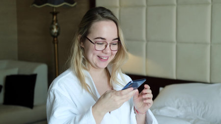 A relaxed woman in a white coat sits on a bed in a hotel room and reads messages on her smartphone....