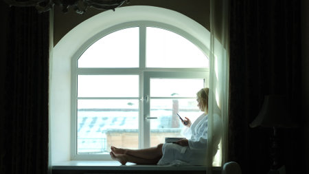 A relaxed woman in a white coat sits on a windowsill in a hotel room and reads messages on her...