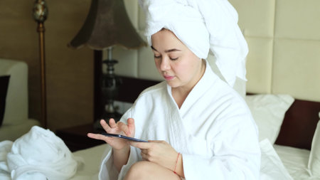 A relaxed woman in a white coat sits on a bed in a hotel room and reads messages on her smartphone. Online communication. Application or messenger.の写真素材