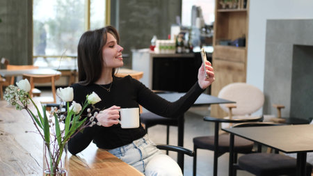 A beautiful girl takes a selfie in a cafe, smiles and looks at the phone. Brunette woman with long hair drinks coffee, rests during lunchの写真素材