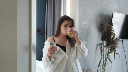 Beautiful woman in a white home shirt looks into her smartphone, drinks water with lemon after waking up in the early morningの写真素材