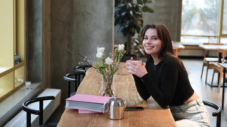 Charming woman with beautiful smile reading good news on mobile phone during rest in coffee shop, happy Caucasian female watching her photo on cell telephone while relaxing in cafe during free timeの写真素材