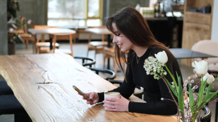 Charming woman with beautiful smile reading good news on mobile phone during rest in coffee shop, happy Caucasian female watching her photo on cell telephone while relaxing in cafe during free timeの写真素材