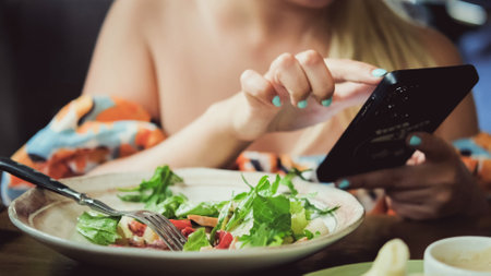 A beautiful blonde woman eats a salad in a cafe during lunch. A business woman uses a smartphone to...