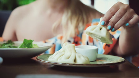 A beautiful blonde woman eats hot khinkali with her hands in a restaurant of traditional Georgian cuisine. Close-up. Khinkali is a national dish of Georgian cuisineの写真素材