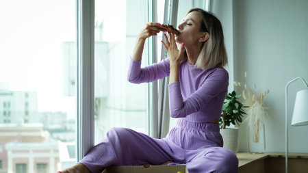 A beautiful lady in a purple home suit sits near the window and eats Italian pizza. Blonde girl enjoys the taste of fresh hot pizzaの写真素材