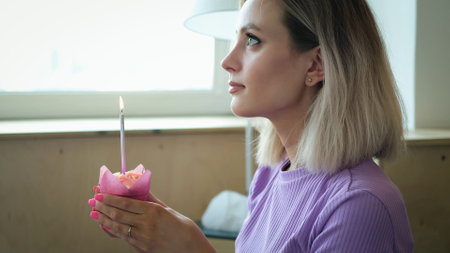 A beautiful young blonde woman wearing a homemade purple suit, holding a festive cupcake with a candle and making a wish. Blonde girl blows out a candle on her birthday cakeの写真素材