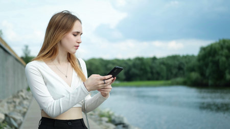 A beautiful young teenage girl in a white top, with long brown hair, uses a smartphone outdoors in the summer, while walking in nature on the shore of a lake. The girl writes text messages and communicates on a social network on her mobile phone.の写真素材