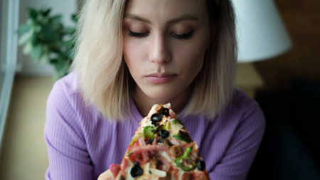A beautiful lady in a purple home suit sits near the window and eats Italian pizza. Blonde girl enjoys the taste of fresh hot pizzaの写真素材
