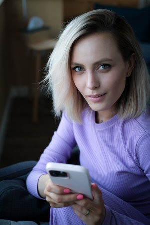 Happy young woman 20 years old in casual clothes, a purple home suit, sitting on a sofa indoors with...