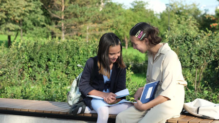 Cute happy little schoolchildren with backpacks and notebooks sitting on a park bench outdoors. Two young schoolgirls doing homework outdoorsの写真素材