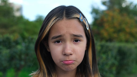 Portrait of a pensive sad little girl, front view, sad child on the street, portrait of an unsmiling schoolgirl, beautiful impassive face of a lonely cute child. The girl lowered her sad eyesの写真素材