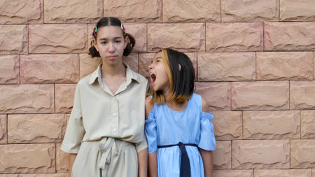 Two cute girls are standing near a peach-colored brick wall. Mixed race sisters of different ages. Teenager and preschooler. Concept of relationship, love in familyの写真素材