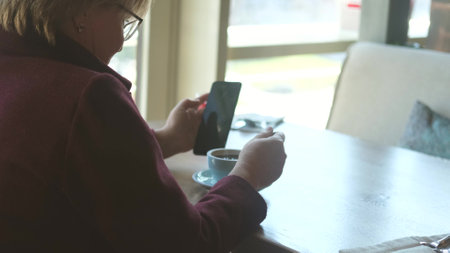 A middle-aged woman with glasses is having lunch in a cafe and reading the news on her smartphone,...