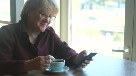 A middle-aged woman with glasses is having lunch in a cafe and reading the news on her smartphone,...