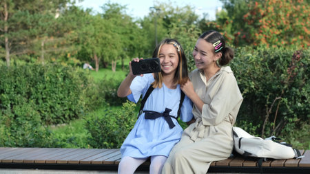 Happy sisters watch a video, take a selfie or talk on a video call using a smartphone outside in the park on a summer day.の写真素材